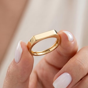 May include: A gold ring with a rectangular top and a polished finish. The ring features a geometric design with faceted sides and a rounded band. The word "FIOR" is engraved on the inside of the ring.