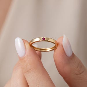 May include: A gold band ring featuring a single, small, round, deep pink gemstone set flush into the metal. The ring has a smooth, polished finish and a simple, circular design, showcasing the vibrant color of the stone against the gold.