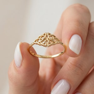 May include: A gold ring featuring a central decorative element composed of two symmetrical, interlocking triquetra knot designs. The interior of the thin, polished gold band is clearly engraved with the brand name FIOR in capital letters.
