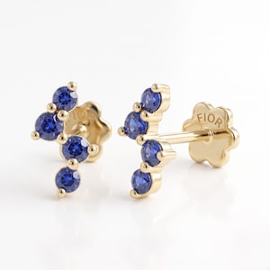 May include: A pair of gold stud earrings featuring a curved design set with four round, vibrant blue stones. The flat, flower-shaped backings are clearly visible, with the word FIOR engraved on the back of each earring post mechanism.