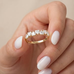May include: A gold ring featuring seven heart-shaped diamonds. The ring is held up by a person with white nail polish. The word "FIOR" is engraved on the ring.