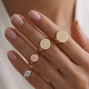 Può includere: Una mano mostra una collezione di anelli sigillo incisi in oro e argento. Gli anelli presentano iniziali personalizzate in uno stile classico ed elegante. Gli anelli sono di varie dimensioni e sono un oggetto di gioielleria popolare.