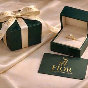 Puede incluir: Un anillo de oro con una piedra clara se encuentra dentro de un joyero verde oscuro abierto. Al lado hay una caja cerrada con una cinta color crema con la palabra FIOR. Una tarjeta verde oscuro muestra un logotipo de ciervo dorado y el texto FIOR LUMIN JEWELLERY.