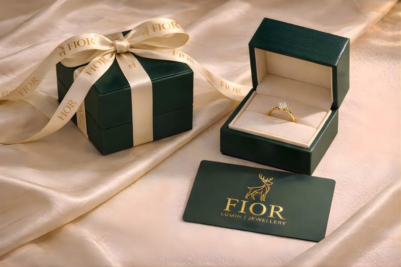Puede incluir: Un anillo de oro con una piedra clara se encuentra dentro de un joyero verde oscuro abierto. Al lado hay una caja cerrada con una cinta color crema con la palabra FIOR. Una tarjeta verde oscuro muestra un logotipo de ciervo dorado y el texto FIOR LUMIN JEWELLERY.