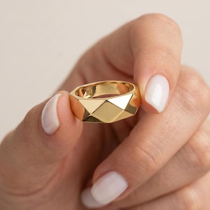 Könnte beinhalten: Ein goldener Bandring mit facettiertem, geometrischem Oberflächendesign. Die Außenseite weist mehrere flache, abgewinkelte Ebenen auf, die ein reflektierendes, mehrseitiges Erscheinungsbild erzeugen. Die Innenseite ist mit FIOR graviert.
