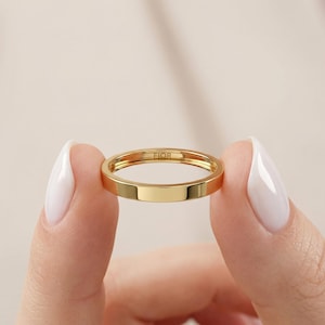 May include: A gold ring with a flat, rectangular band is held between two fingers. The ring has the word "FIOR" engraved on the inside. The ring is a simple, elegant design.