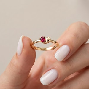 May include: A gold ring with a heart-shaped design. It features a teardrop-shaped red gemstone and a heart-shaped accent. The band is engraved with "FIOR". The ring is held up by a person.
