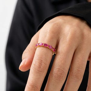 Op de afbeelding: Een gouden ring met een rij ovaal geslepen, helder roze edelstenen. De ring is gezet in een geelgouden band, wat een contrast cre&euml;ert met de heldere edelstenen. De ring wordt getoond op een vinger.