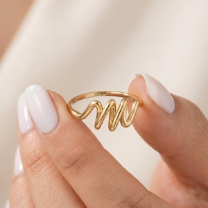May include: A gold ring with a distinctive, flowing design. The ring has the word "FIOR" inscribed on the band. The ring is held between two fingers with white nail polish.