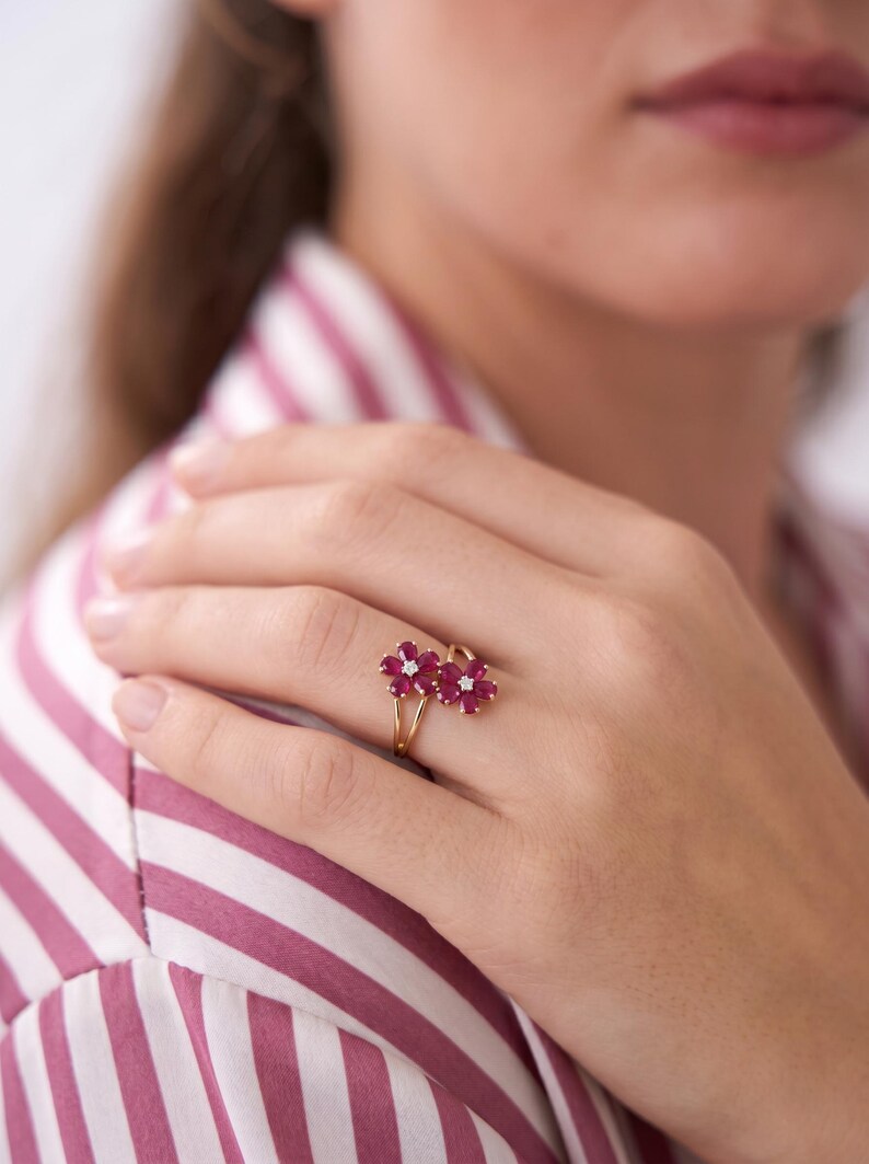 Puede incluir: Un anillo de oro con dos motivos en forma de flor, cada uno compuesto por cinco rub&iacute;es de color rosa intenso en talla marquesa que rodean un peque&ntilde;o diamante blanco central. El aro es fino y pulido, con los dos elementos florales juntos.