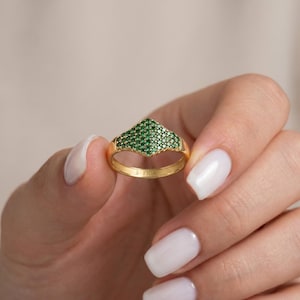 Könnte beinhalten: Ein Goldring mit einem schildförmigen Kopf, der mit zahlreichen kleinen, runden, leuchtend grünen Steinen im Pavé-Stil besetzt ist. Die Ringschiene ist glatt und poliert, wobei das Wort FIOR deutlich auf der Innenseite des Goldrings eingraviert ist.