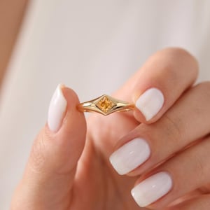 Può includere: Un anello in oro con una singola pietra preziosa gialla a taglio quadrato incastonata in una montatura a forma di diamante. La fascia è liscia e lucida, si restringe leggermente verso l'incastonatura centrale che sostiene la pietra.