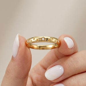 May include: A gold ring featuring star and diamond details. The ring has a smooth finish and the word "FIOR" is engraved on the band. The ring is held between two fingers.