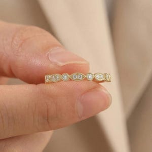May include: A delicate gold ring with a repeating pattern of small, sparkling diamonds. The ring's design alternates between oval and round diamond settings, creating a continuous, elegant band. The ring is held by a hand against a soft, neutral background.