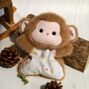 May include: A plush monkey toy with brown faux fur, a light pink face, and black eyes. The monkey wears a white fabric garment featuring a pattern of small, multicoloured stars in shades of yellow, orange, pink, and blue across the front.