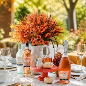 Op de afbeelding: Een witte keramieken kan gevuld met kunstmatige oranje en gele bloemen en varens, geplaatst op een tafel met twee wijnflessen, twee wijnglazen, een kleine oranje taartstandaard en drie kleine witte en grijze decoratieve pompoenen.