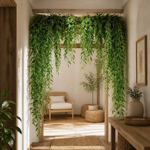 May include: Two long, cascading artificial willow vine garlands with vibrant green leaves hanging from a wooden ceiling beam, creating a lush, dense curtain of foliage that frames the entrance to a bright, open interior space.