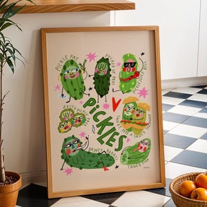 Pode incluir: Uma impressão artística emoldurada com picles de desenho animado com várias expressões e nomes como "Pickle Pat" e "Briney Badass". A palavra "Pickles" é proeminente. Fundo marrom claro, moldura de madeira.