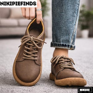 Op de afbeelding: Bruine leren veterschoenen met een ronde neus en een dikke, lichtbruine zool. De schoenen hebben lichtbruine veters en een gele binnenvoering. De afbeelding toont de schoenen die worden vastgehouden door een persoon die een blauwe spijkerbroek draagt.