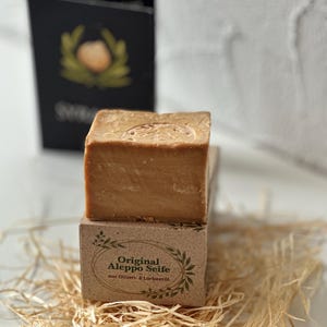 Könnte beinhalten: Ein hellbraunes Aleppo-Seifenstück liegt auf einem Karton mit dem Text "Original Aleppo Seife aus Oliven- & Lorbeeröl". Die Seife wird auf Stroh präsentiert, mit einer schwarzen Karte im Hintergrund. Ein Naturprodukt für die Haut.