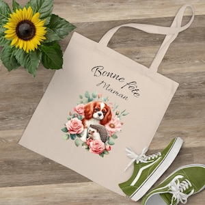 May include: Beige tote bag with a floral design featuring a Cavalier King Charles Spaniel and a hedgehog, surrounded by pink roses. The text "Bonne fête Maman" is printed on the bag. A green sneaker is visible in the image.