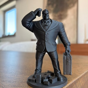 Op de afbeelding: Een zwarte 3D-geprinte figuur van een man in een pak, met een aktetas. De figuur staat op een ronde basis met stapels blokken. De man draagt een pak en stropdas, met zijn rechterhand naar zijn hoofd geheven.