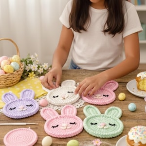 Op de afbeelding: Gehaakte konijnvormige onderzetters in pastelkleuren: lavendel, wit, roze en groen. Elke onderzetter heeft geborduurde ogen en een neus. Een haaknaald en een mand met paaseieren zijn ook zichtbaar.