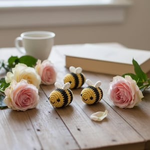 Può includere: Tre api all'uncinetto fatte a mano con strisce gialle e nere e ali bianche sono disposte su un tavolo di legno. Rose rosa e crema e una tazza di tè sono anche sul tavolo, insieme a un libro.