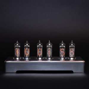 May include: A nixie tube clock featuring six glass vacuum tubes mounted on a rectangular silver-colored metal base. The tubes display glowing orange numerical digits, specifically showing the sequence 1, 0, 0, 1, 5, 8, against a dark background.