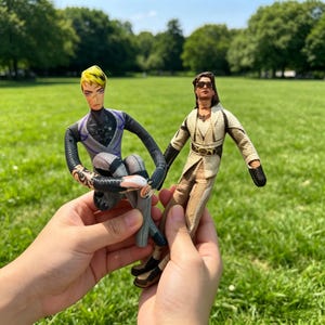 May include: Two fabric dolls are held up against a green grassy background. One doll is wearing a black and grey outfit with a purple vest and yellow hair. The other doll is wearing a tan outfit with black accents and sunglasses.