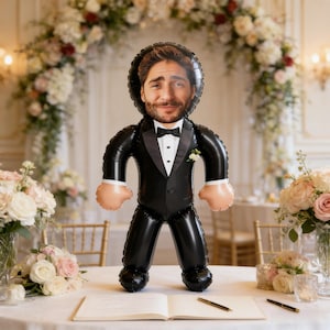 Könnte beinhalten: Ein schwarzer Ballon in Form eines Mannes im Smoking, mit aufgedrucktem Gesicht. Der Ballon hat hautfarbene Hände und eine weiße Blume am Revers. Der Hintergrund zeigt einen Hochzeitsbogen und Blumengestecke.