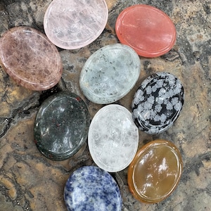 May include: Assortment of oval-shaped gemstones in various colors, including pink, red, gray, green, white, black, blue, and amber. The stones are arranged on a textured, brown surface.