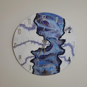 Può includere: Orologio da parete rotondo con sfondo bianco e design astratto blu e viola. L'orologio presenta numeri e lancette argentate. Il design ricorda un fiume o un ruscello.