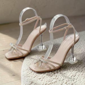 Pu&ograve; includere: Un paio di eleganti sandali con tacco alto. Le scarpe presentano un plantare color nudo, tacchi trasparenti e sottili cinturini. Un dettaglio a fiocco scintillante adorna il cinturino della punta. I sandali sono esposti su un tappeto dai toni neutri.