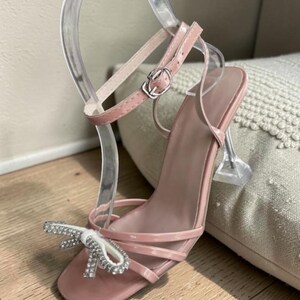 Pu&ograve; includere: Un sandalo con tacco alto rosa cipria con tacco trasparente e curvo e punta quadrata. Il sandalo presenta un delicato cinturino alla caviglia con fibbia argentata e un fiocco impreziosito da strass. La scarpa &egrave; esposta su un espositore trasparente.