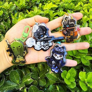 May include: Five acrylic keychains depicting cartoon characters in tactical gear. The keychains feature characters with weapons, helmets, and distinct designs. Each keychain has a different colour scheme and is attached to a metal ring or clasp.