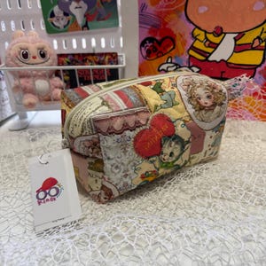 Op de afbeelding: Een gewatteerde make-uptas met een patchwork-ontwerp met vintage illustraties, waaronder een meisje, een kat en harten. De tas heeft een ritssluiting en een label met een logo. Het kleurenpalet omvat crème, roze, rood en geel.