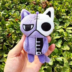 Puede incluir: Un peluche de gato en tonos morados y blancos, con un diseño cosido. El gato tiene un ojo negro y una boca cosida. El juguete se sostiene en una mano sobre un fondo de follaje verde.