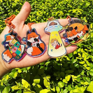 Könnte beinhalten: Vier Acryl-Schlüsselanhänger mit bunten Illustrationen. Einer zeigt einen Fuchs in einer Patchwork-Decke, ein anderer einen Fuchs mit Brille, ein dritter eine Figur, die von einem UFO entführt wird mit dem Text AMAZE, und der letzte einen Fuchs, der einen Burger mit der Aufschrift ME BURGER isst.