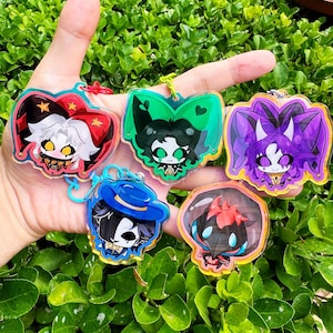 May include: Five acrylic charms featuring stylised cartoon characters. One has a red jester hat, one is green with cat ears, one is purple with horns, one wears a blue hat, and one is a dark bird-like figure. Each charm has a small metal attachment.