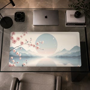 Puede incluir: Una alfombrilla de escritorio rectangular con una ilustración de un paisaje que muestra un lago tranquilo rodeado de montañas bajo un gran sol pálido. Una rama de cerezo con flores rosas se extiende desde la izquierda sobre la escena azul claro.