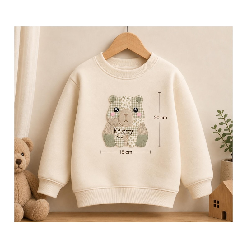 Puede incluir: Una sudadera de manga larga color crema con un gr&aacute;fico central de oso con patr&oacute;n de patchwork. El dise&ntilde;o del oso incluye el texto "Nizzy" y "Nest" con l&iacute;neas de medici&oacute;n visibles que indican una altura de 20 cm y una anchura de 18 cm.