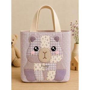 Puede incluir: Un bolso tote de color morado claro con un dise&ntilde;o de oso de patchwork en la parte delantera. El oso est&aacute; confeccionado con varios patrones de tela, incluyendo cuadros vichy, flores y espiga. El nombre Nizzy est&aacute; bordado en el pecho.