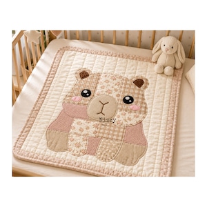 Puede incluir: Una manta de beb&eacute; acolchada cuadrada con un dise&ntilde;o de oso de patchwork en tonos beige, crema y rosa suave. El oso tiene ojos negros bordados, mejillas rosadas y el texto Nizzy impreso en el pecho. Un peluche de conejo se encuentra cerca.