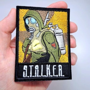 Pode incluir: Patch bordado com uma figura em máscara de gás e equipamento de proteção, com uma mochila e um rifle. O patch tem uma borda preta, um fundo amarelo e marrom, e a palavra "STALKER" em letras brancas.