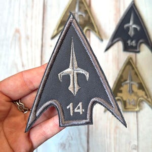 以下が含まれることがあります： 矢印型のパッチのコレクション。メインのパッチはグレーで、茶色の縁取り、茶色のエンブレム、数字の14が付いています。背景にある他のパッチは、ゴールドとブラックの色合いです。
