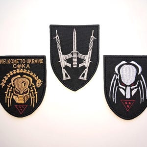 Puede incluir: Tres parches negros bordados con varios diseños. Uno presenta el texto "WELCOME TO UKRAINE C#KA" sobre una criatura dorada. Otro muestra una espada y rifles. El tercero muestra una criatura blanca y roja.