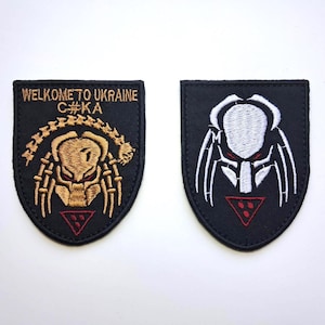 Puede incluir: Dos parches negros bordados. Uno presenta un diseño tipo Predator con el texto "WELCOME TO UKRAINE C#KA" en dorado y rojo. El otro parche tiene un diseño Predator blanco con detalles en rojo.
