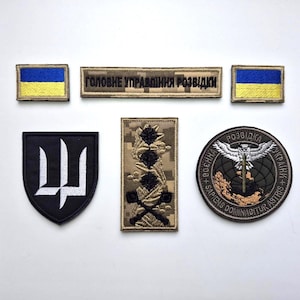 Puede incluir: Un conjunto de parches bordados con símbolos militares y gubernamentales ucranianos. Incluye parches con la bandera ucraniana, un tridente y texto en ucraniano. Los parches varían en forma y diseño, con una mezcla de colores.