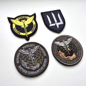 Op de afbeelding: Vier geborduurde patches met militaire ontwerpen. De patches bevatten Oekraïense en Latijnse tekst, samen met symbolen zoals een drietand en een gestileerde adelaar. De kleuren zijn zwart, geel, wit en olijfgroen.